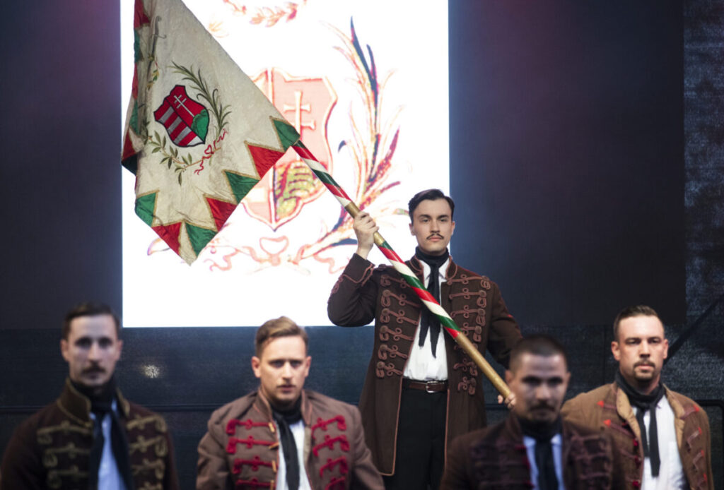 Férfiak korhű, díszmagyar viseletben állnak a színpadon, a szabadsághős előadás jelenetében. A középen álló férfi magasra emelt magyar zászlót tart.