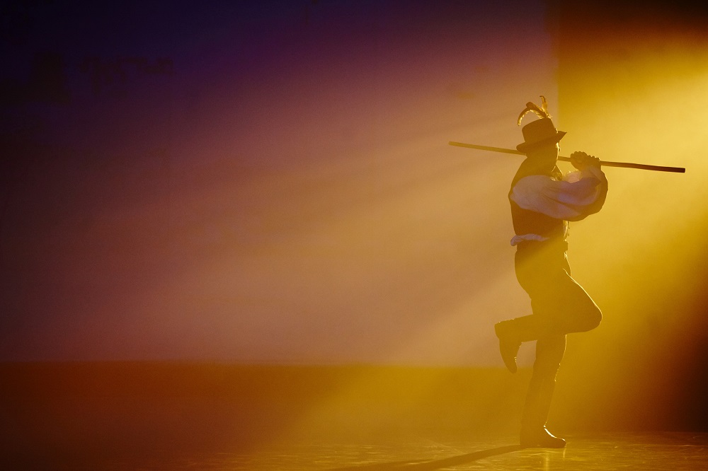 Puszták népe előadás, férfi táncos puskával a vállán, hagyományos viseletben, sárgás fényben megvilágított színpadon.