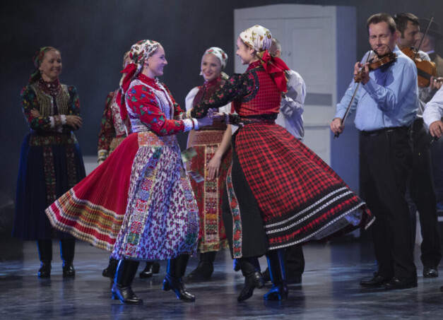 Népviseletbe öltözött táncosok és zenészek előadása Székesfehérváron, két nő forgatós párostáncot táncol, hegedűs zenészek kíséretével.