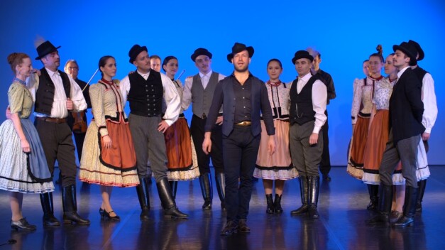 Kis Magyar Táncrajz előadás. Népviseletbe öltözött férfiak és nők állnak a színpadon kék háttér előtt, középen egy férfi énekel.